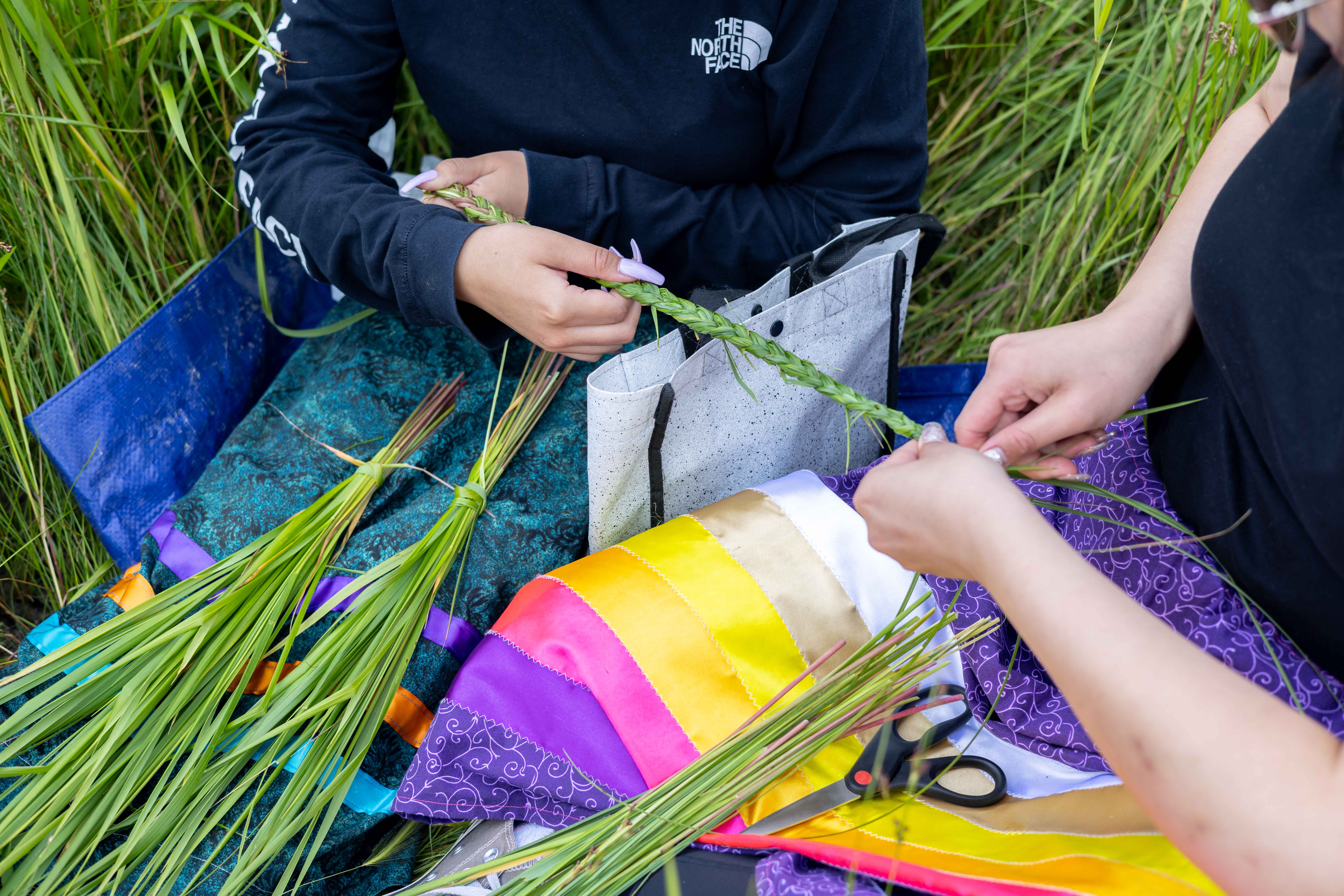 Two people are braiding sweetgrass while seated on the ground. 